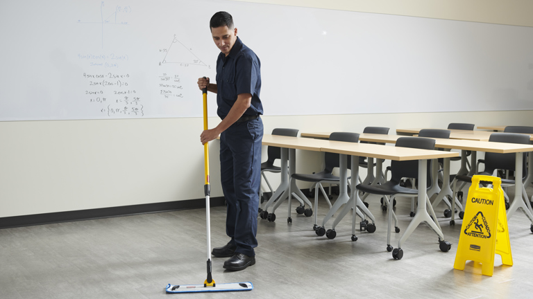 Male custodian cleaning the floor of an empty classroom with a flat mop, with furniture visible in the background.