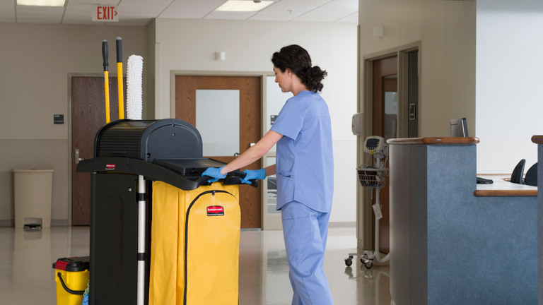 Person in blue scrubs and gloves pushing a Rubbermaid High Security Cleaning Cart with yellow bag and tools in a medical hallway.