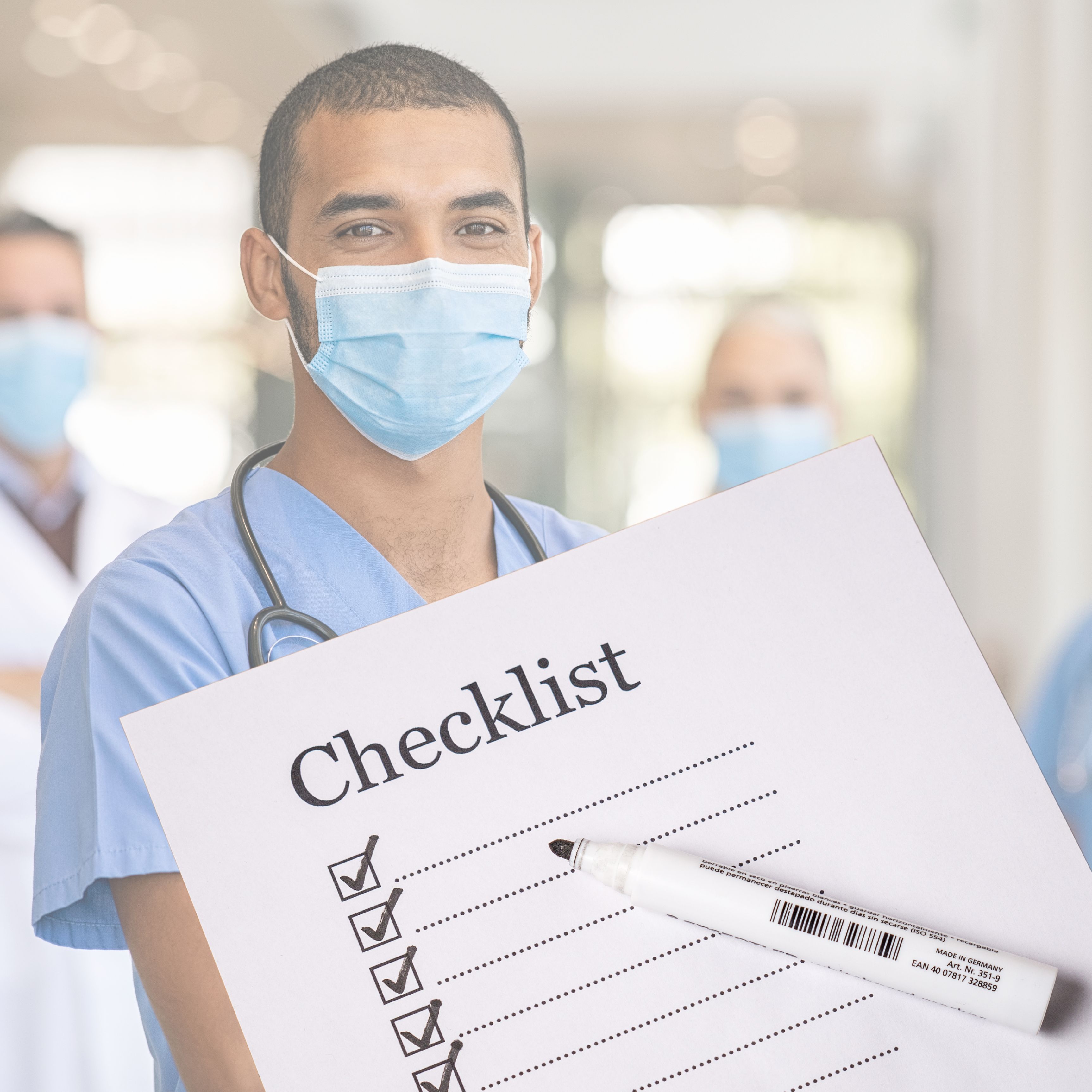 Male healthcare professional in blue scrubs and face mask holding a checklist with first four items ticked, stethoscope around his neck, in a bright hospital setting.
