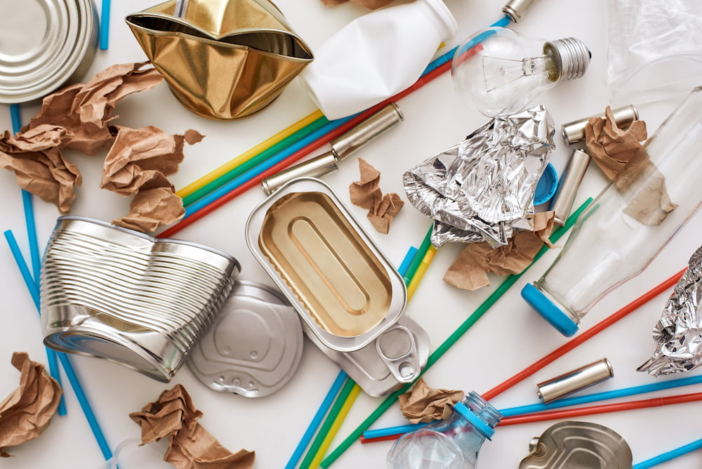 Assorted recyclable waste including crumpled aluminum and tin cans, glass and plastic bottles, a light bulb, and plastic straws on a white surface.